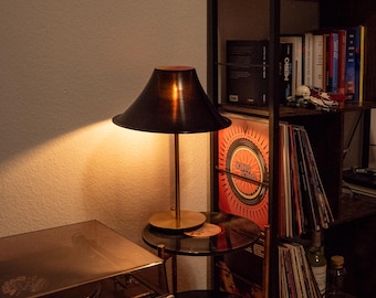 May include: A black table lamp with a copper-colored base and shade sits on a two-tiered side table. The side table holds vinyl records. A record player is to the left, and a bookshelf with records and books is to the right.