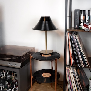 Op de afbeelding: Een unieke bijzettafel gemaakt van gerecyclede vinylplaten, met twee niveaus en koperkleurige poten. Bovenop staat een zwarte lamp met een messing steel en kap. De tafel staat naast een plank met vinylplaten en boeken.