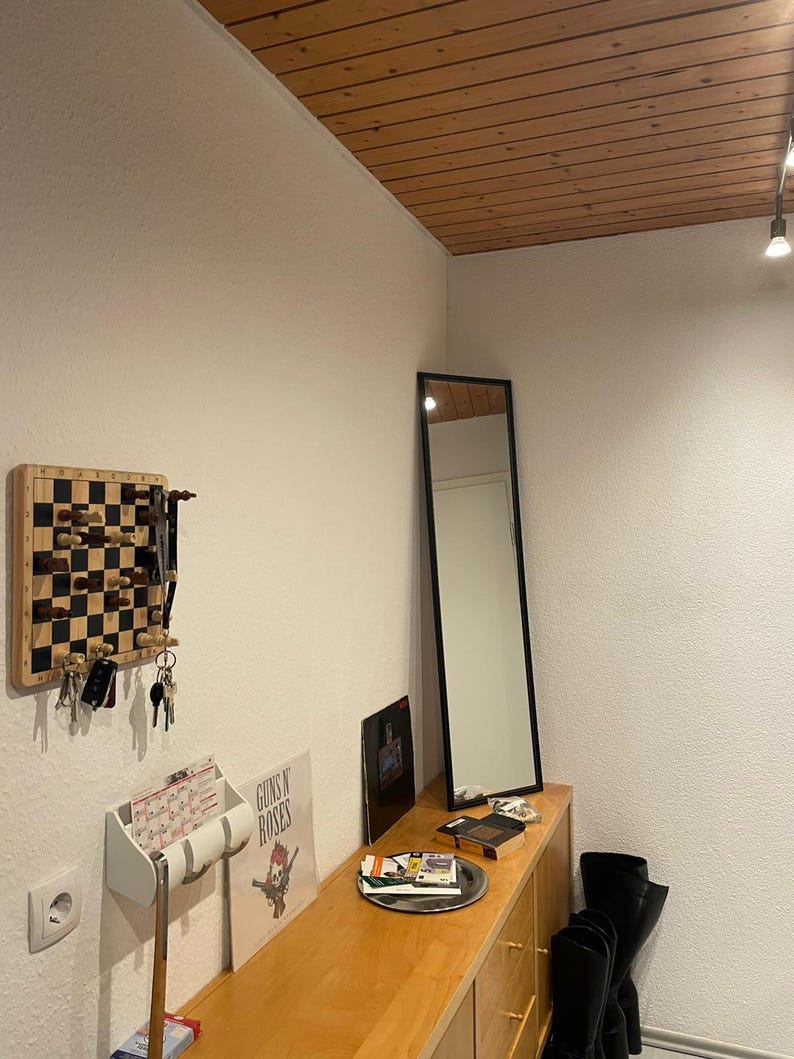 May include: A wooden entryway with a white wall, a wooden ceiling, a black mirror, a wooden shelf with a Guns N' Roses album, a white key holder, and a chessboard with chess pieces.