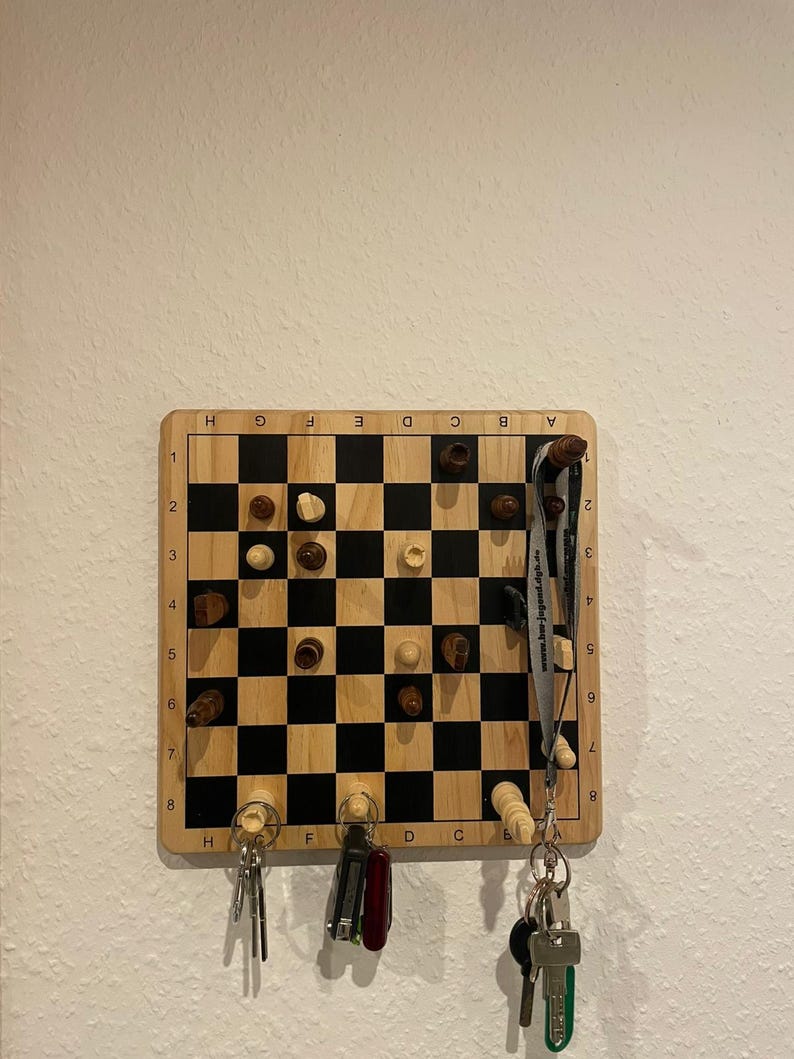 May include: A wooden magnetic chessboard with white and black squares, with chess pieces on the board. The board is mounted on a wall and has key hooks on the bottom. There are keys and a pocket knife hanging on the hooks.
