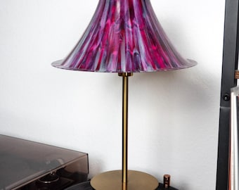 May include: A table lamp with a brass base and stem, topped with a lampshade featuring a vibrant pattern of pink, purple, and gray. The lamp sits on a black vinyl record, creating a retro aesthetic.