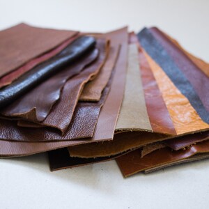 May include: A collection of leather swatches in various shades of brown, tan, and burgundy. The textures vary from smooth to textured, with some pieces folded and layered. The lighting highlights the different tones and finishes of the leather.