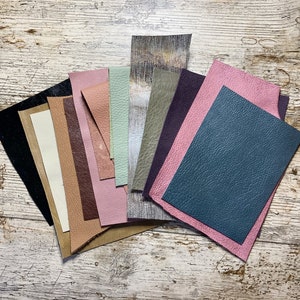 May include: A collection of leather swatches in various colors, including black, cream, brown, pink, green, and blue. The leather pieces have different textures and finishes, arranged on a distressed, white-washed wooden surface. Ideal for crafting or design projects.