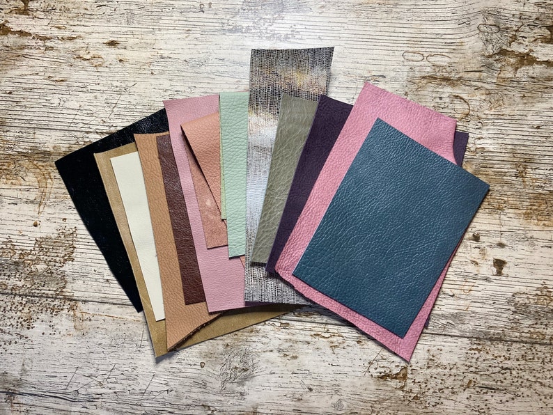 May include: A collection of leather swatches in various colors, including black, cream, brown, pink, green, and blue. The leather pieces have different textures and finishes, arranged on a distressed, white-washed wooden surface. Ideal for crafting or design projects.