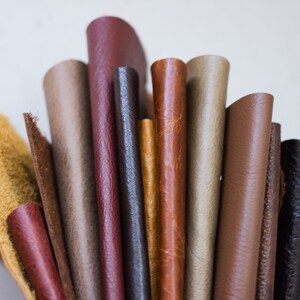 May include: A collection of rolled leather samples in various shades of brown, tan, and burgundy. The textures vary from smooth to slightly textured, showcasing the different finishes and grains available. The image highlights the natural colors and variations of the leather.