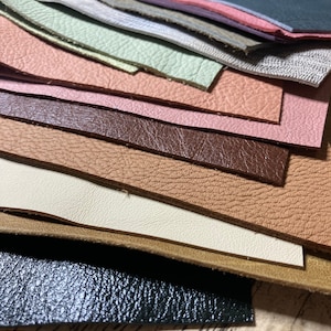 May include: A collection of leather swatches in various colors and textures. The swatches include shades of pink, brown, green, and gray, with different grain patterns. The leather pieces are arranged in a layered, overlapping manner, showcasing the variety of materials.