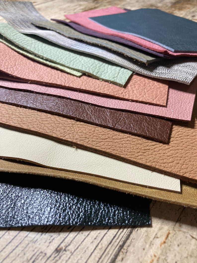 May include: A collection of leather swatches in various colors and textures. The swatches include shades of pink, brown, green, and gray, with different grain patterns. The leather pieces are arranged in a layered, overlapping manner, showcasing the variety of materials.