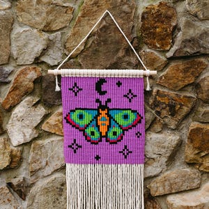 May include: Purple and white macrame wall hanging with a butterfly and crescent moon design. The butterfly is green, orange, and yellow. The wall hanging has a fringe at the bottom.