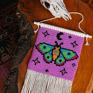 May include: Purple and white wall hanging with a beaded butterfly design and a crescent moon. The butterfly has orange, green, and blue accents. The hanging has a fringe of white yarn.
