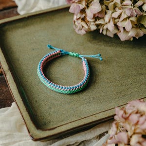 May include: A woven friendship bracelet with pink, green, and blue threads. The bracelet is on a green ceramic plate.