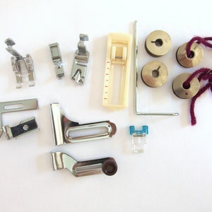 May include: A collection of sewing machine accessories, including various presser feet, bobbins, and a needle plate. The presser feet are silver-toned, and the bobbins are metallic. A small, clear plastic foot with a blue accent is also included. A length of purple thread is also present.