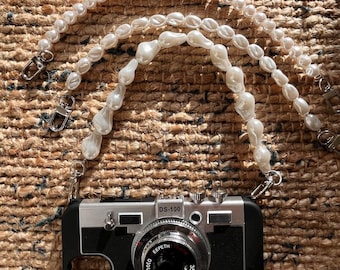 May include: A white pearl camera strap with a silver and black camera case with the text "DS-100" and "1-Photo EEFETH 35mm 1:3.5" on the front.