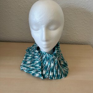 Puede incluir: Un cuello tejido en tonos turquesa, blanco y gris, exhibido en una cabeza de maniqu&iacute; blanca. El cuello est&aacute; hecho de un hilo texturizado y parece suave y c&aacute;lido. El dise&ntilde;o es un patr&oacute;n de punto acanalado simple.