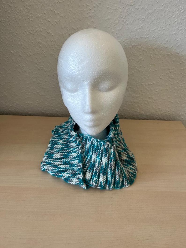 Puede incluir: Un cuello tejido en tonos turquesa, blanco y gris, exhibido en una cabeza de maniqu&iacute; blanca. El cuello est&aacute; hecho de un hilo texturizado y parece suave y c&aacute;lido. El dise&ntilde;o es un patr&oacute;n de punto acanalado simple.