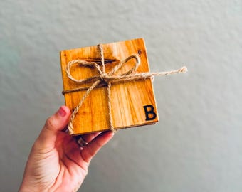 Puede incluir: Un juego de cuatro posavasos de madera atados con cuerda. Los posavasos son de color marrón claro y tienen una veta de madera natural. La letra "B" está quemada en el posavasos superior.