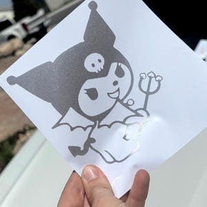 May include: A silver glitter vinyl sticker depicting a cartoon character with a skull on its head and a trident. The character has bat wings and is smiling. The sticker is on a white background.