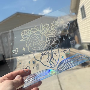 May include: Clear, holographic sticker with a floral heart design, including roses, leaves, and a heart outline. Additional heart and star accents are included. The bottom of the sticker has a layered, iridescent design.