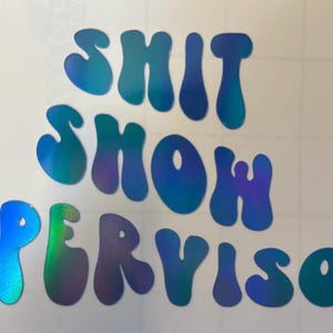 Op de afbeelding: Een holografische sticker met de woorden "SHIT SHOW SUPERVISOR" in een bubbellettertype. De tekst verloopt in tinten blauw, groen en paars. De sticker staat op een witte achtergrond met een rasterpatroon.