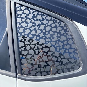 May include: A car window with a decorative film featuring a pattern of gray, irregular shapes resembling cow spots. The window is framed by black trim and is part of a white vehicle.
