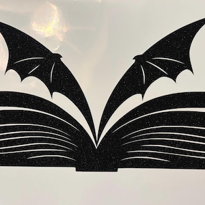 Puede incluir: Silueta negra de un libro abierto con alas de murciélago que se extienden desde la parte superior de cada página. Varias formas de estrella están dispersas alrededor de las alas, creando un tema mágico y de fantasía. El diseño está sobre un fondo claro.