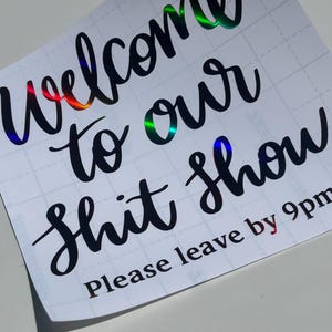 Puede incluir: Adhesivo blanco con texto negro en cursiva que dice: "Welcome to our shit show". El texto tiene un brillo holográfico. Debajo, dice "Please leave by 9pm".