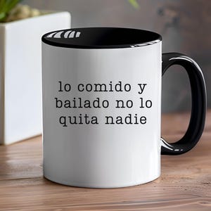 May include: White ceramic mug with a black interior and handle. The mug features the text "lo comido y bailado no lo quita nadie" in black, centered on the front. The mug is on a wooden surface.