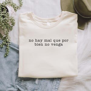 May include: A cream coloured t-shirt with the Spanish phrase "no hay mal que por bien no venga" printed in black.