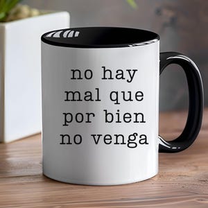 May include: White ceramic mug with a black interior and handle. The mug features the Spanish text "no hay mal que por bien no venga" in black, centered on the front. The mug is on a wooden surface.