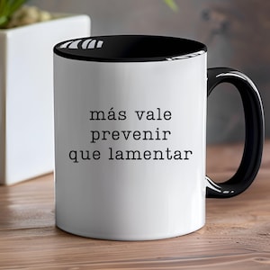 May include: White ceramic mug with a black interior and handle. The mug features the Spanish phrase "más vale prevenir que lamentar" in black text. The mug is on a wooden surface.