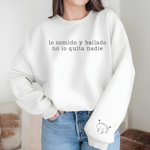 May include: White sweatshirt with the text "lo comido y bailado no lo quita nadie" in black. A small cartoon taco graphic is on the left sleeve. The sweatshirt is paired with blue jeans.