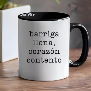 May include: A white ceramic mug with a black interior and handle. The mug features the Spanish text "barriga llena, corazón contento" in black, centered on the front. The mug is on a wooden surface.