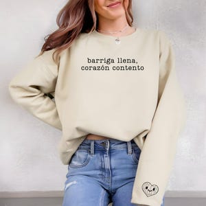 May include: A light beige sweatshirt with the text "barriga llena, corazón contento" printed on the front. The sweatshirt has a small heart graphic on the left sleeve.