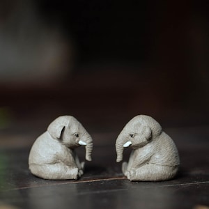 May include: Two small gray ceramic elephant figurines sitting facing each other on a dark wooden surface.