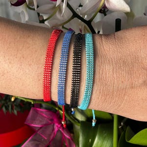 Puede incluir: Cuatro pulseras coloridas con cuentas plateadas, cada una atada con un nudo y una pequeña cuenta dorada en el extremo. Las pulseras son roja, azul, negra y turquesa.