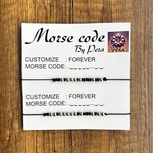 May include: Two black and silver beaded bracelets with black string. The bracelets are on a white card with the text "Morse code by Pera" and "CUSTOMIZE: FOREVER" and the Morse code for "FOREVER".