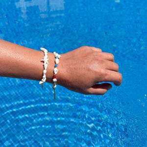 May include: Two seashell bracelets. One bracelet features white starfish and shells. The other bracelet has white shells, red, blue, and green beads. Both bracelets are on a wrist, with a blue water background.