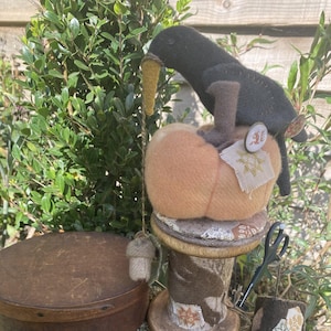 Könnte beinhalten: Eine handgefertigte Herbstdekoration mit einer Filzkrähe auf einem Kürbis, einer Holzspule, einer runden Holzkiste und einem Gusseisenstern. Die Krähe hält eine gelbe Maiskolben. Der Kürbis sitzt auf einer Spule mit Stoffdesign.