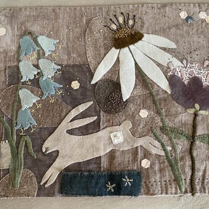 May include: Textile artwork depicting a running rabbit, bluebell flowers, and a large white daisy. The piece uses brown, green, and white tones, with a patchwork background. Decorative floral elements are scattered across the surface of the artwork.