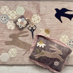 May include: A pink fabric sewing kit featuring a bunny and flower design. The kit includes a small pillow with a bunny applique, scissors, and various patterned hexagon fabric pieces. A dark blue bird silhouette is also present.
