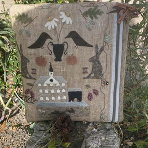 May include: A rectangular beige fabric pouch featuring intricate needlework embroidery of a house, two rabbits, two black birds, a vase with white flowers, and pumpkins. The pouch has a white zipper closure on the right side with a brown ribbon tie.