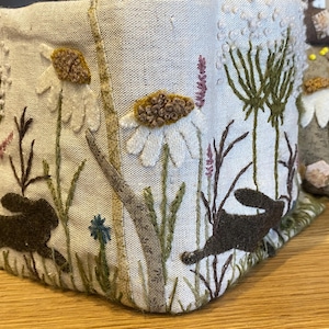 May include: A fabric item with embroidered details. The design features white daisies, brown rabbits, and various wildflowers in shades of green, brown, and purple. The item is placed on a wooden surface.