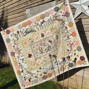Peut inclure: Une courtepointe faite à la main sur le thème de la nature, avec des lapins, des fleurs et des motifs hexagonaux aux couleurs pastel douces. La courtepointe est exposée à l'extérieur, mettant en valeur son design et son savoir-faire complexes. La courtepointe est en tissu.