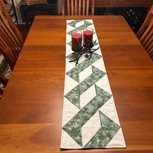 May include: A rectangular table runner with a geometric pattern of green and white shapes, adorned with red candles in glass holders. The runner features a repeating design of angled blocks with snowflake details, placed on a wooden table.