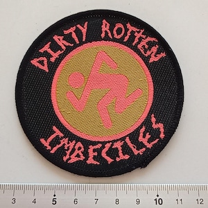 May include: A black and pink round patch with the text "DIRTY ROTTON" in black and "IMBECILES" in pink around a gold circle with a pink stick figure inside.
