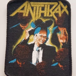 Op de afbeelding: Een zwarte geweven patch met een gele band aan de bovenkant waarop in hoofdletters "ANTHRAX" staat. De patch toont een kleurrijk beeld van een man in pak en hoed, met een gele en blauwe achtergrond.