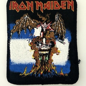 Op de afbeelding: Iron Maiden band patch met een zwarte achtergrond, een rood-wit-blauwe vlag en een gevleugelde schedel met vlammen.