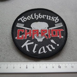 Op de afbeelding: Zwarte ronde patch met de tekst "Toothbrush Chariot Klan!" in wit en rood. Het ontwerp bevat gekruiste tandenborstels en een schedel. De patch heeft een diameter van ongeveer 7,6 cm.
