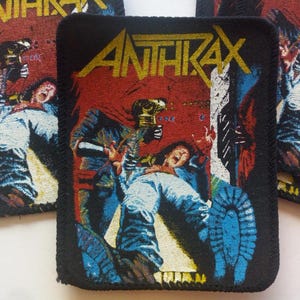 May include: Anthrax band patch with a black border. The patch features the band's name in yellow block letters over a red background. The image depicts a scene with figures in blue and white clothing, with a distressed, vintage aesthetic.