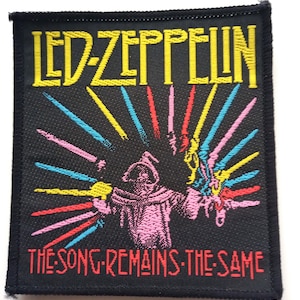 Op de afbeelding: Zwarte geweven patch met de tekst "LED ZEPPELIN" in geel en "THE SONG REMAINS THE SAME" in rood. Een roze figuur met een kap en meerdere kleurrijke zwaarden bevindt zich in het midden.