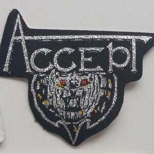 Op de afbeelding: Een zwart en zilver geborduurde patch met het woord "ACCEPT" in zilveren letters en een zilveren en zwarte schedel met rode ogen.